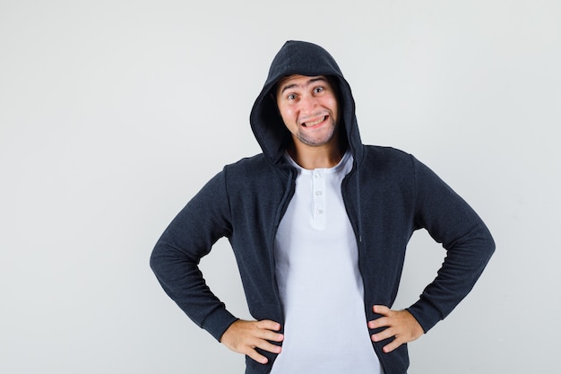 Young male holding hands on waist in jacket, t-shirt and looking cheerful. front view.