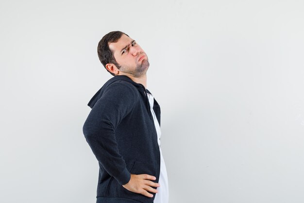 Young male holding hands on waist in t-shirt, jacket and looking bored. .