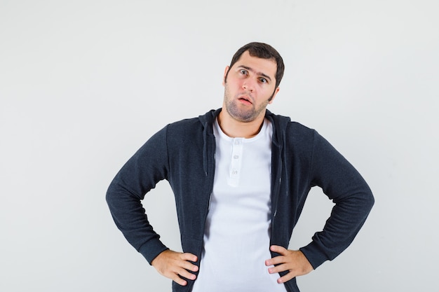 Young male holding hands on waist in t-shirt, jacket and looking confused , front view.