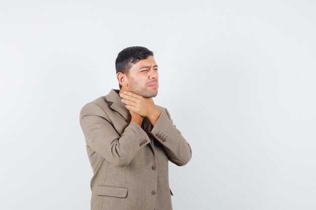 Young male holding his throat in grayish brown jacket and looking sick , front view.