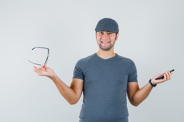 Free Photo young male holding mobile phone and glasses in grey t-shirt cap and looking jolly 