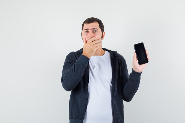 Young male holding mobile phone in t-shirt, jacket and looking surprised. front view.