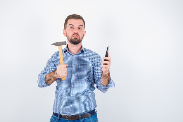 Young male holding mobile phone while keeping hammer in shirt, jeans and looking serious. front view.