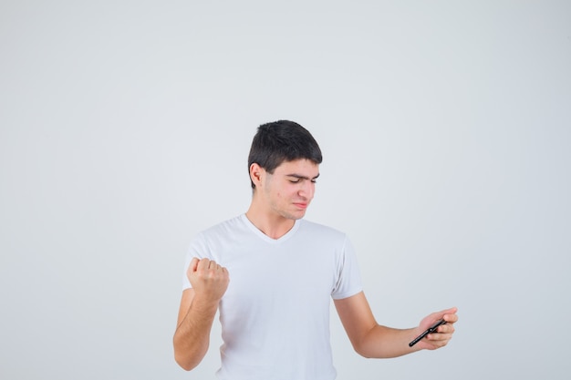 Young male holding phone, showing clenched fist in t-shirt and looking cheery. front view.