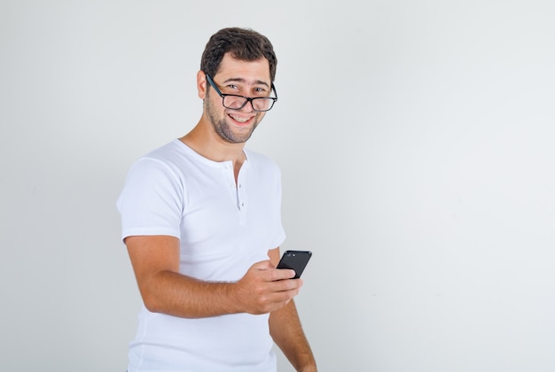 Young male holding smartphone and laughing in white t-shirt, glasses and looking merry