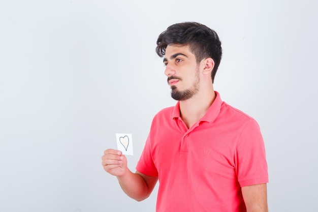 Young male holding sticky note while looking away in t-shirt and looking thoughtful , front view.