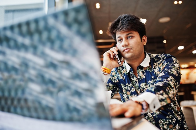 Free photo young male indian freelancer sitting open laptop computer in fast food cafe handsome asian man speaking phone during work on netbook in comfortable coffee shop