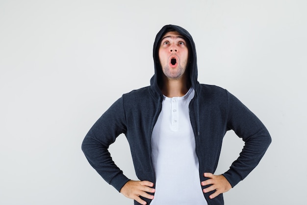 Young male in jacket, t-shirt holding hands on waist and looking amazed , front view.