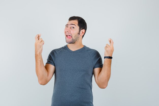 Young male keeping fingers crossed winking eye sticking out tongue in grey t-shirt 