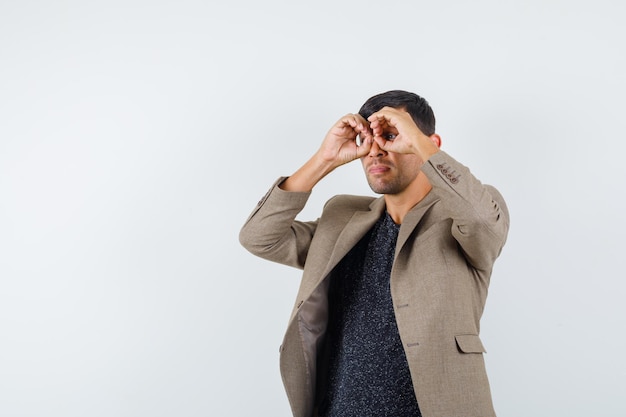 Young male looking away while showing glasses gesture with his fingers in grayish brown jacket,black shirt and looking weird .