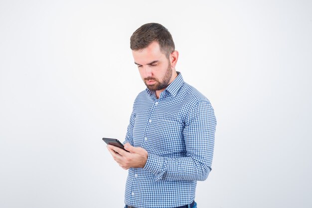 Young male looking at mobile phone in shirt, jeans and looking serious. front view.