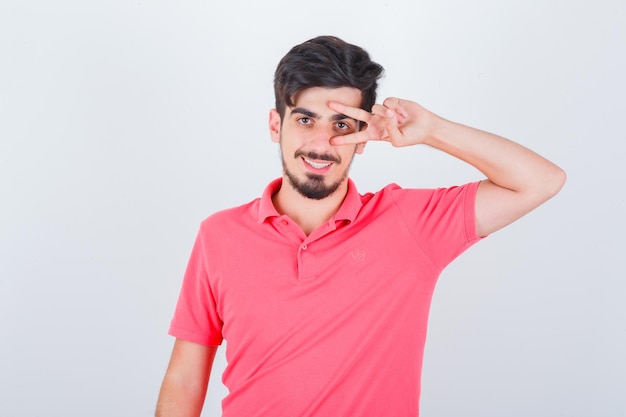 Young male in pink t-shirt showing V-sign on eye and looking cheerful , front view.