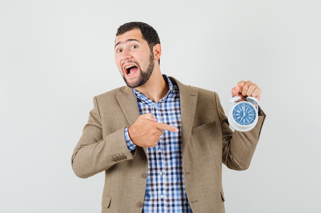 Young male pointing at alarm clock in shirt, jacket and looking happy. front view.