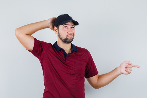 Young male pointing aside while looking away in red t-shirt,black cap and looking pensive , front view.