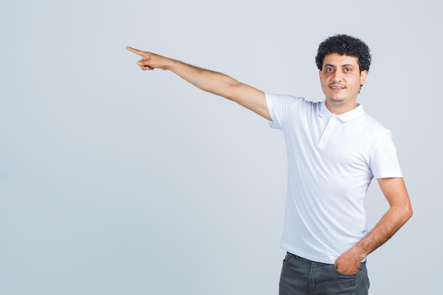 Young male pointing aside in white t-shirt, pants and looking confident. front view.