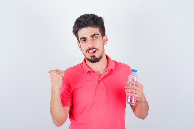 Young male pointing back with thumb while sticking out tongue in t-shirt and looking happy , front view.