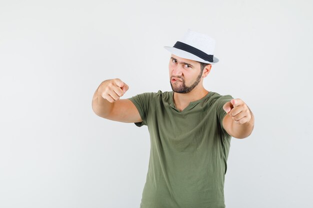 Young male pointing at camera, biting lip in green t-shirt and hat and looking focused