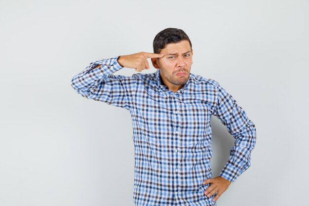 Young male pointing finger at temples in checked shirt and looking upset