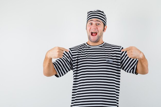 Young male pointing at himself in t-shirt, hat and looking merry. front view.