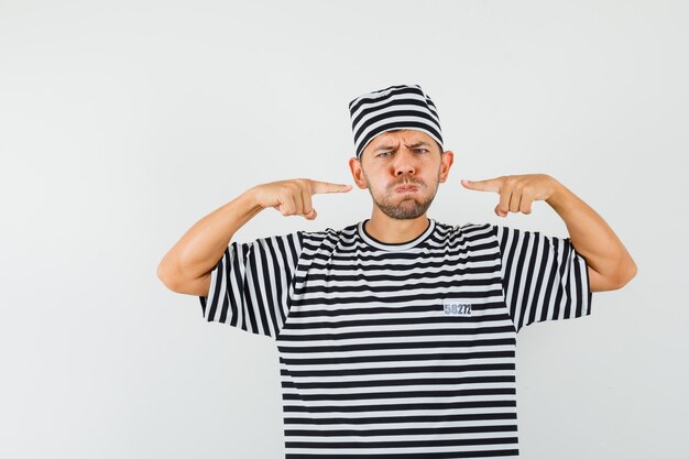 Young male pointing at his blown cheeks in striped t-shirt hat and looking gloomy 