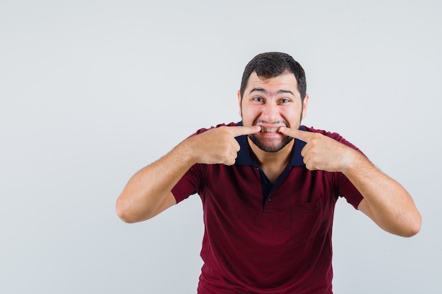 Young male pointing at his teeth in red t-shirt , front view.