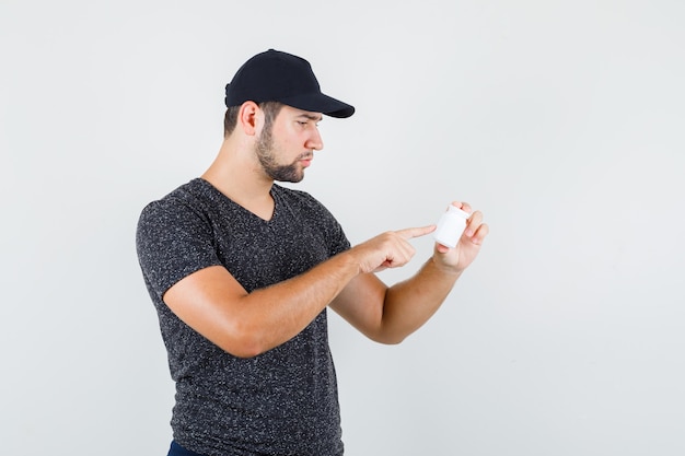 Young male pointing at info on bottle of pills in t-shirt and cap and looking careful