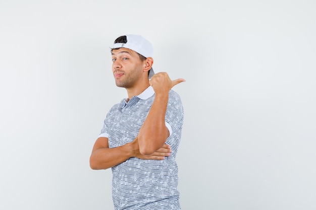 Young male pointing thumb away in t-shirt and cap and looking cunning