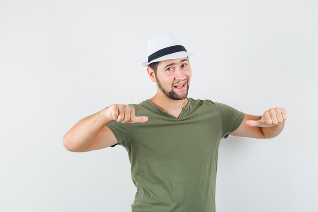 Young male pointing thumbs at himself in green t-shirt and hat and looking cool