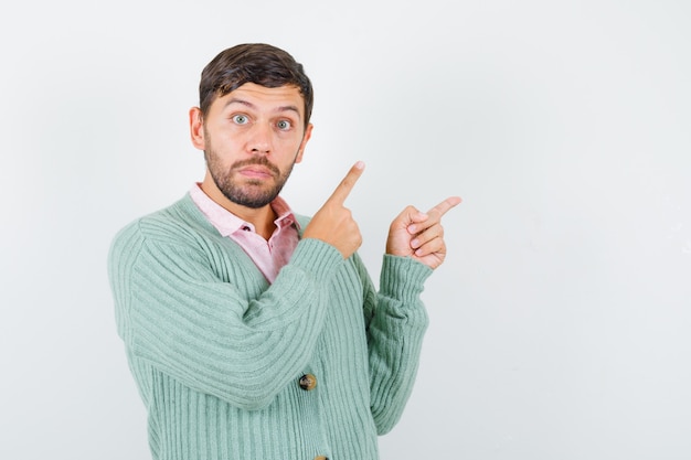 Free photo young male pointing at upper right corner in shirt, cardigan and looking puzzled , front view.