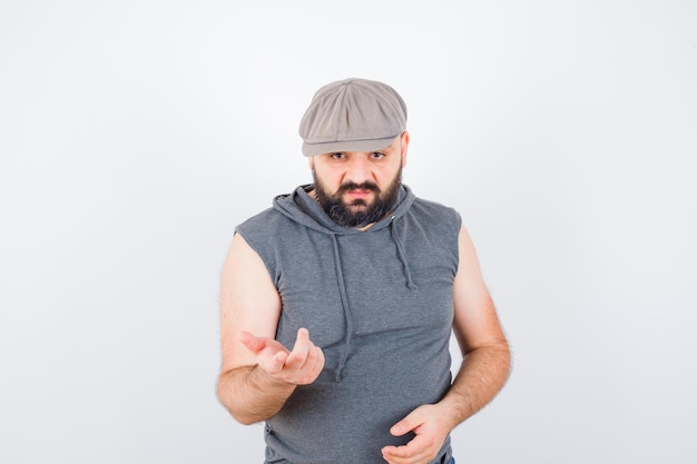 Young male raising hand in questioning pose in sleeveless hoodie, cap and looking serious , front view.