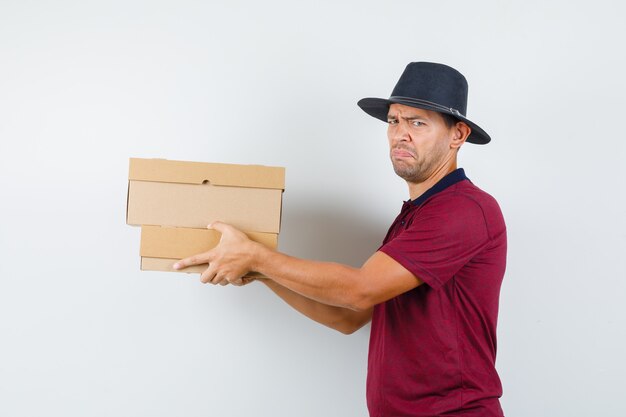 Young male in red shirt,black hat carrying boxes and looking displeased .