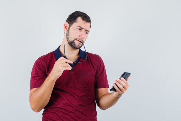 Young male in red t-shirt looking at phone and looking puzzled , front view.