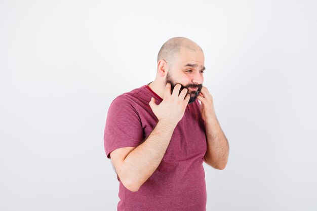 Young male scratching his beard in pink t-shirt and looking troubled , front view.