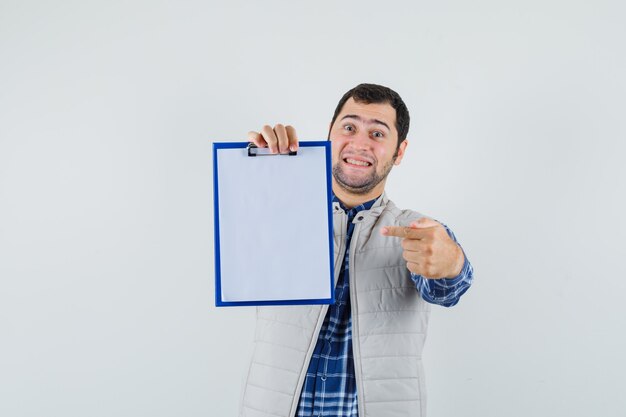 Young male in shirt,jacket pointing to blank clipboard and looking contented , front view.