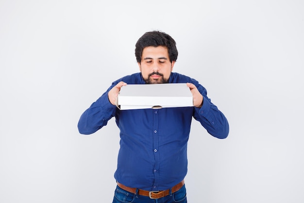 Young male in shirt, jeans holding closed pizza box and looking pretty , front view.