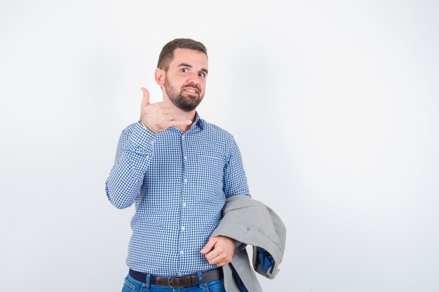 Young male in shirt, jeans, suit jacket holding suit jacket while showing phone gesture and looking happy , front view.