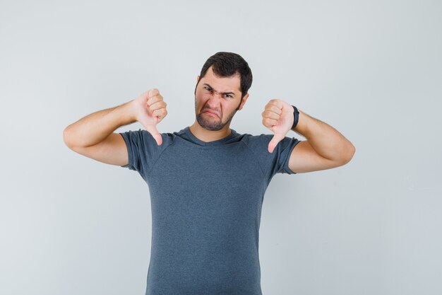 Young male showing double thumbs down in grey t-shirt and looking downcast. front view.