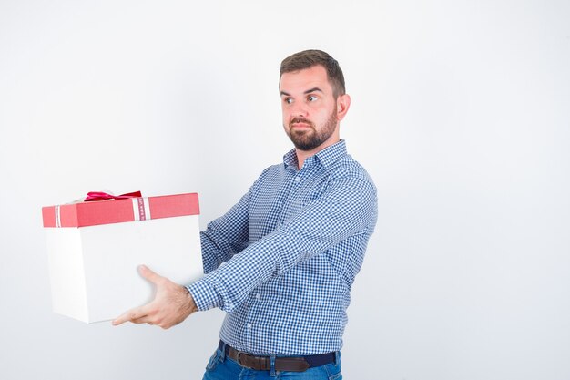 Young male showing giving gesture while holding gift box in shirt, jeans and looking confident , front view.