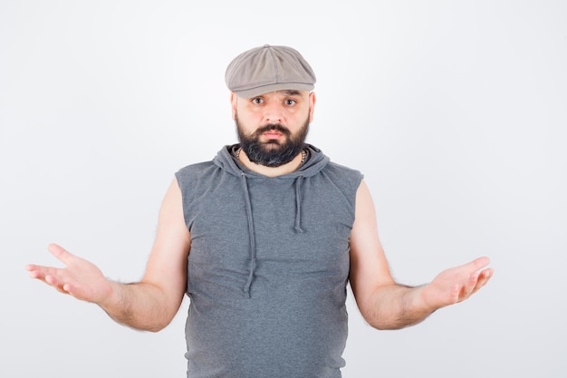 Young male showing helpless gesture in sleeveless hoodie, cap and looking desperate. front view.