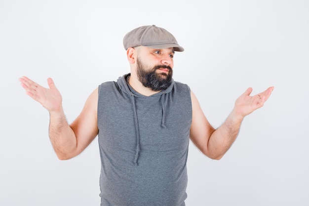 Free photo young male showing helpless gesture in sleeveless hoodie, cap and looking desperate. front view.