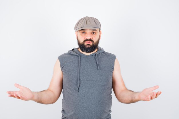 Young male showing helpless gesture in sleeveless hoodie, cap and looking wistful. front view.