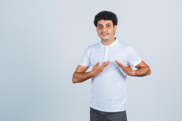 Young male showing herself in questioning gesture in white t-shirt, pants and looking proud. front view.