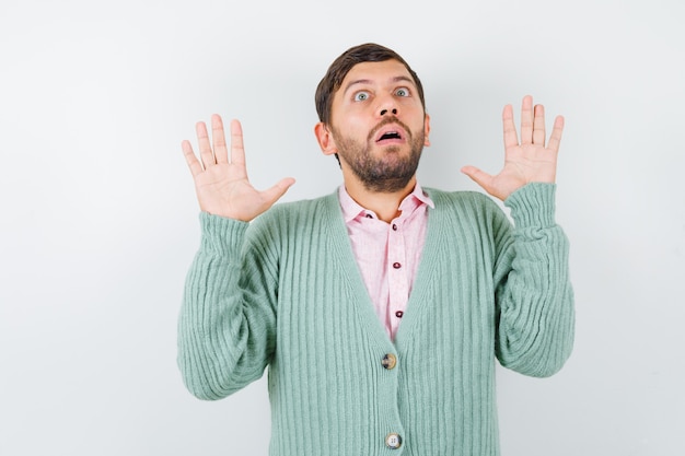 Young male showing palms in surrender gesture in shirt, cardigan and looking scared. front view.