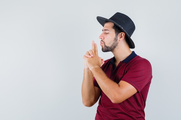 Young male showing pistol sign in t-shirt,hat and looking confident , front view.