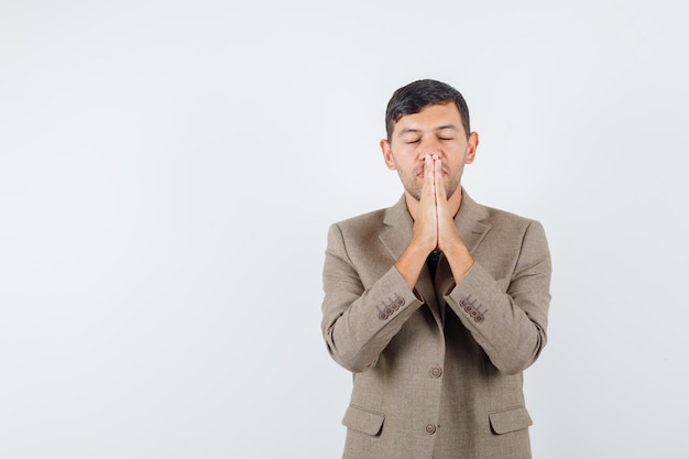 Young male showing praying gesture in grayish brown jacket and looking wishful. front view. free space for your text