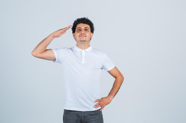 Young male showing salute gesture in white t-shirt, pants and looking confident , front view.