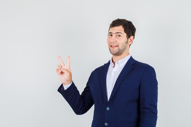 Young male showing victory sign in suit and looking joyful. front view.