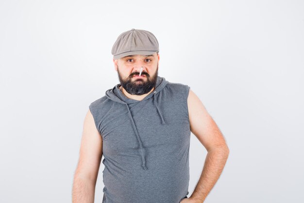 Young male in sleeveless hoodie, cap posing while standing and looking confident , front view.