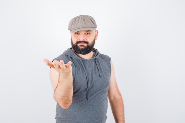 Young male in sleeveless hoodie, cap raising hand in questioning pose and looking confident , front view.