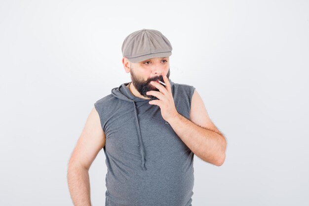 Young male in sleeveless hoodie, cap smoking while posing and looking confident , front view.